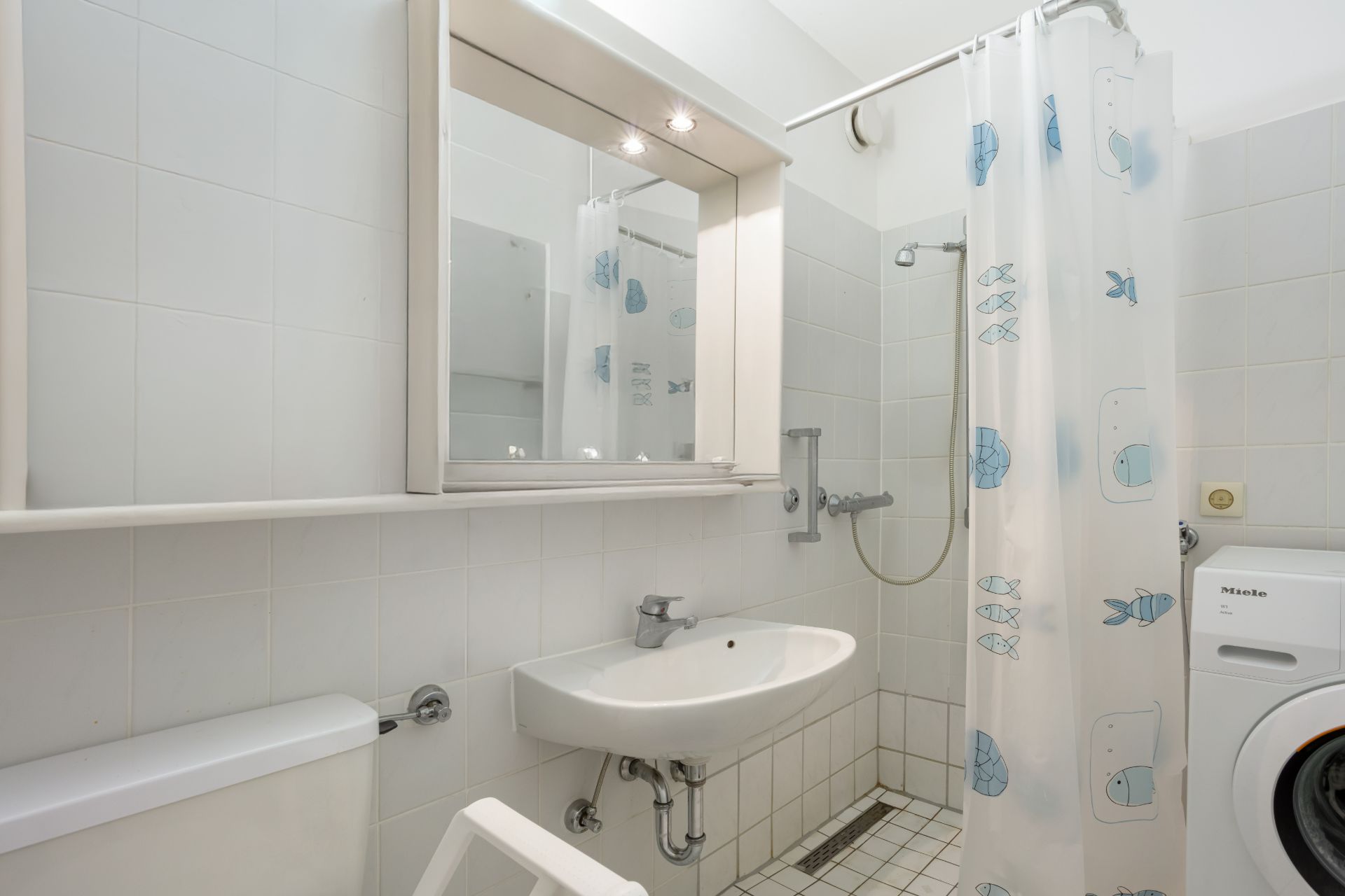 Helles Badezimmer mit Waschbecken, Spiegel, Toilette, Dusche mit Duschvorhang und Waschmaschine.