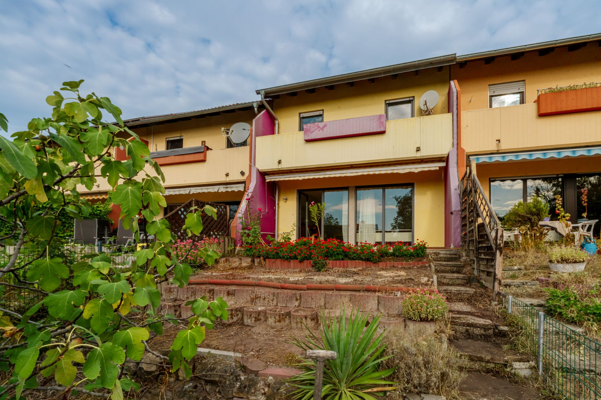 Zweistöckiges Wohnhaus mit Balkon, Terrasse, Garten und Außentreppe, im Vordergrund Pflanzen.