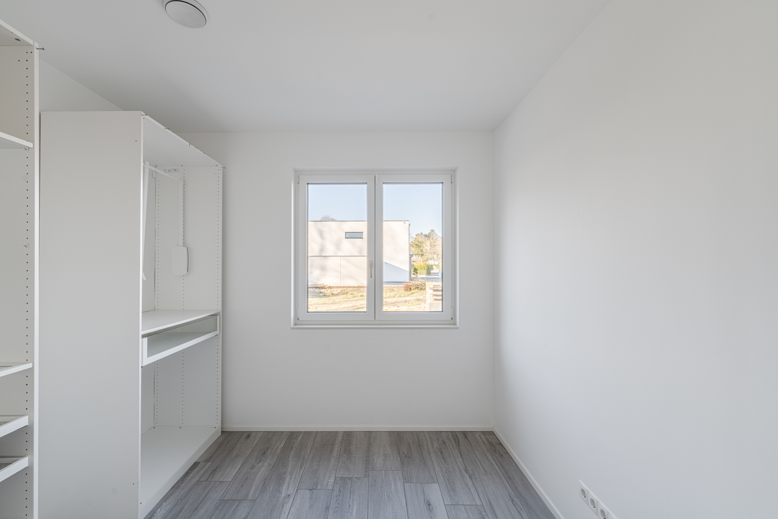 Heller, leerer Raum mit Fenster, grauem Boden und weißem Einbauschrank links.