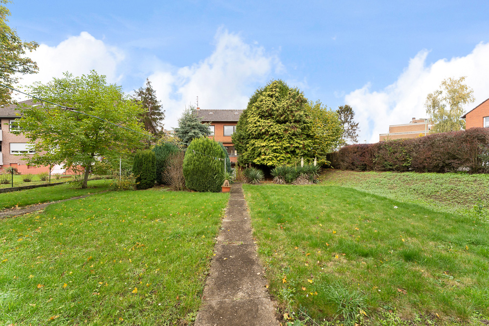 Garten mit Rasenfläche, Gehweg und verschiedenen Bäumen, Hecken und Sträuchern im Hintergrund.