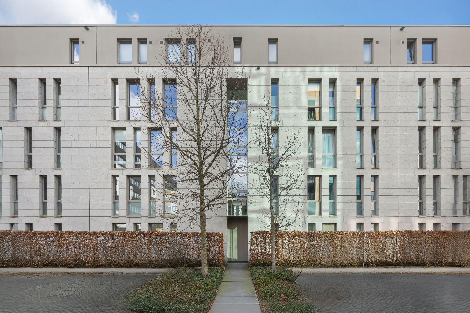 Moderne Wohnhausfassade mit vielen Fenstern, zwei Bäumen und Hecke im Vordergrund.
