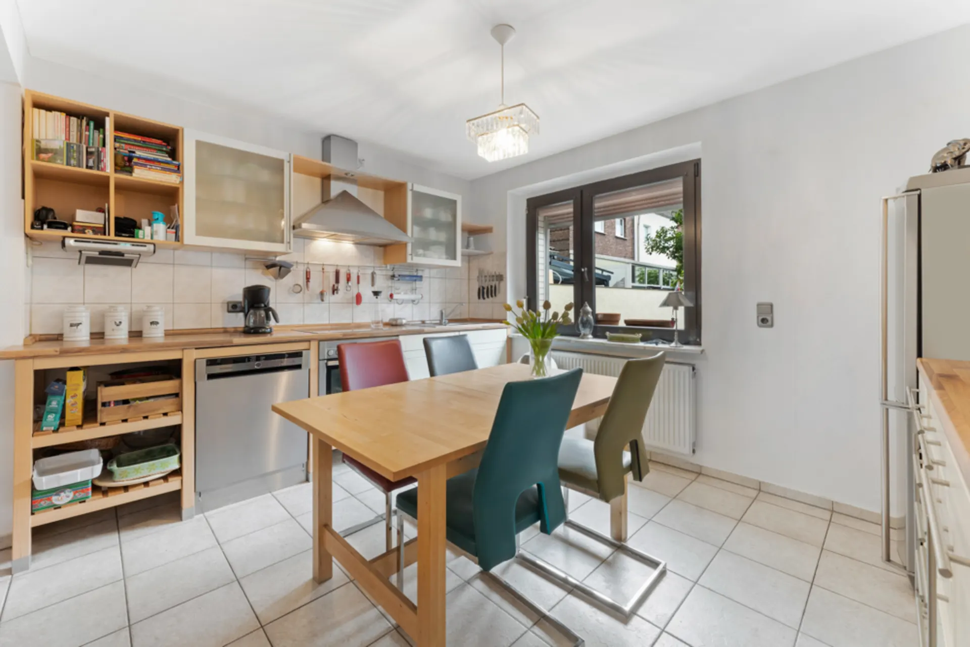 Helle Küche mit Holztisch, bunten Stühlen und großem Fenster, moderne Geräte und offene Regale.