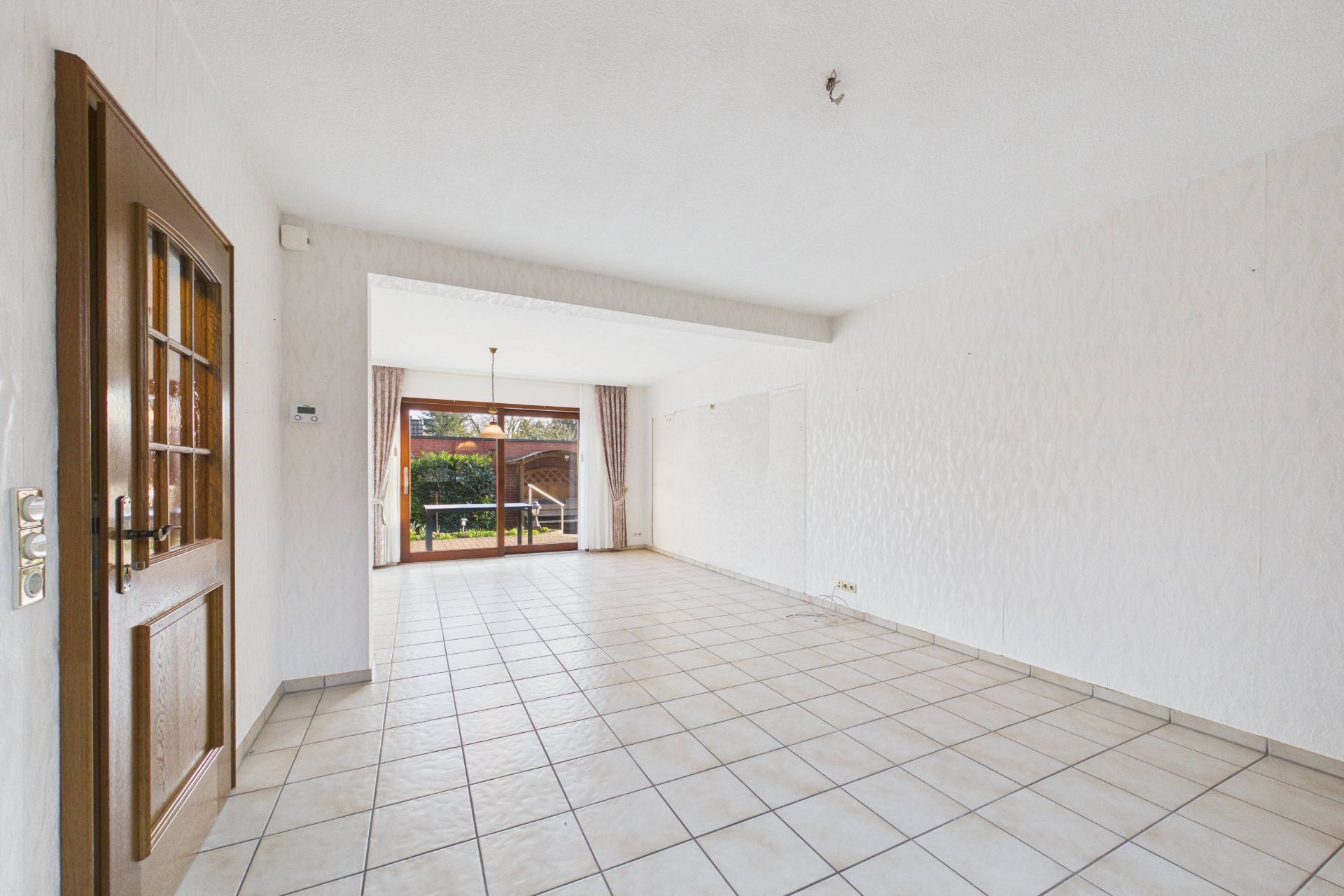 Helles, leeres Wohnzimmer mit Fliesenboden und großer Glastür zum Garten.