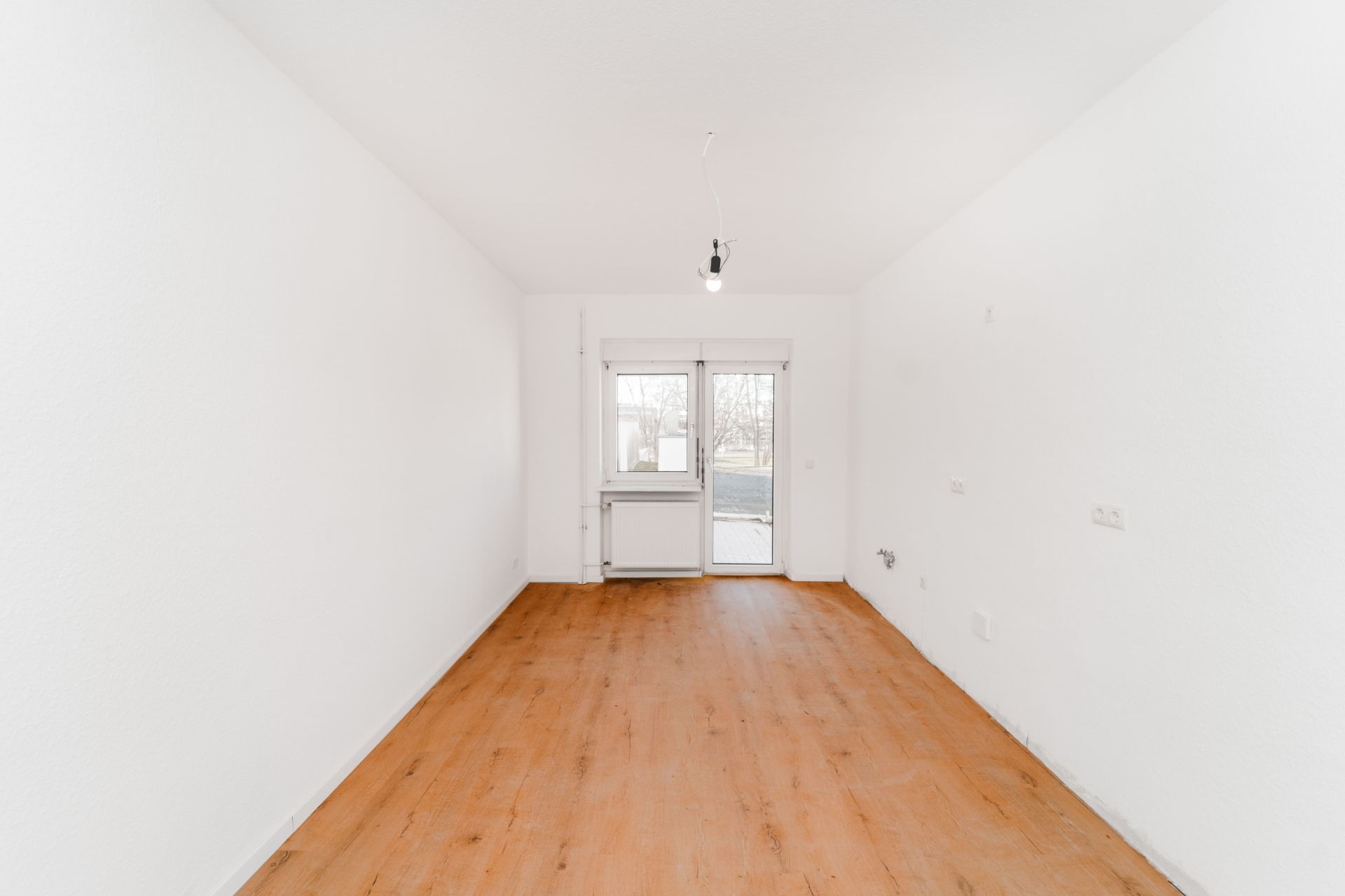 Leerer Raum mit weiß gestrichenen Wänden, Holzboden und Fenster mit Tür zum Balkon.