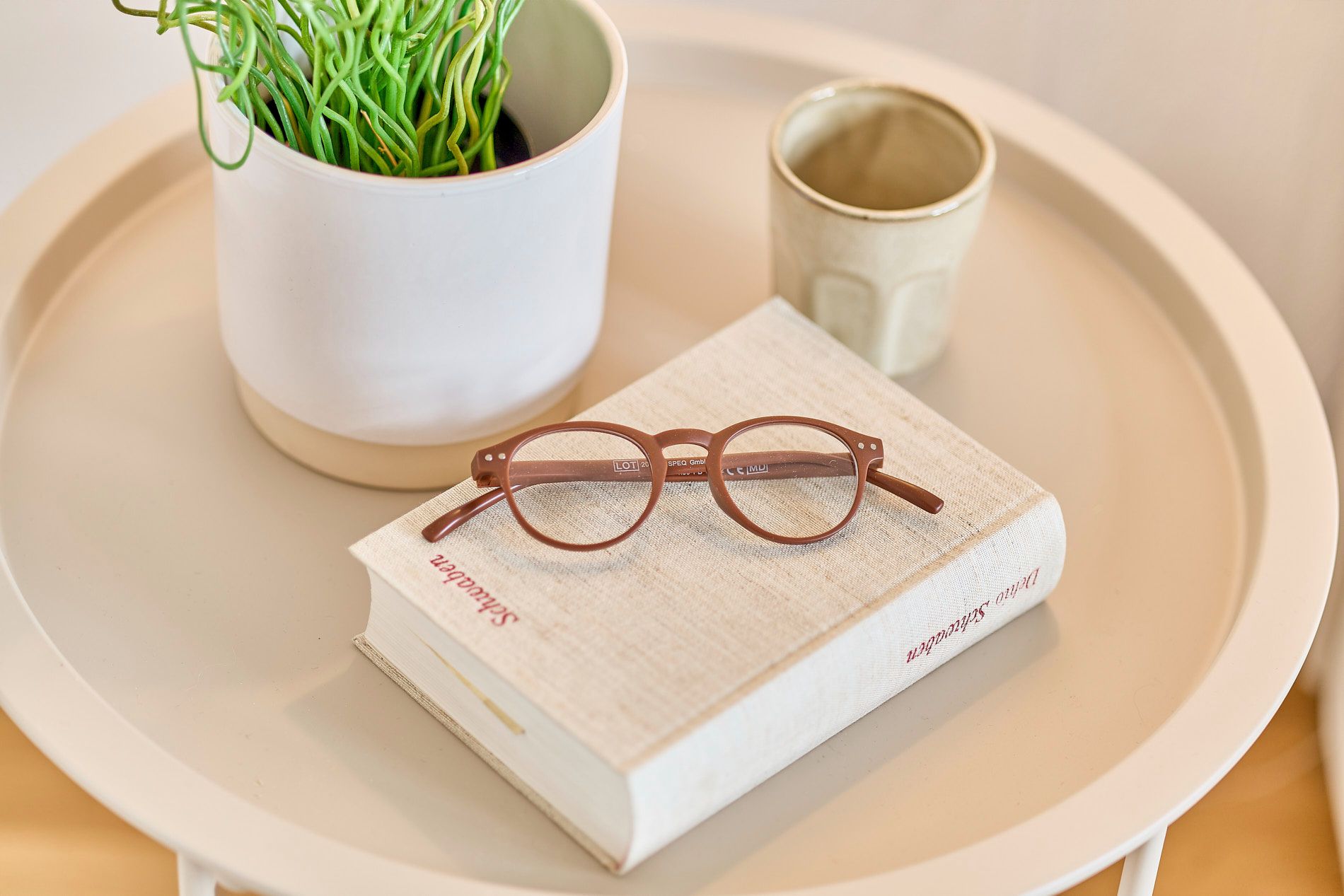 Brille auf Buch neben Pflanze und Becher auf rundem Tisch.