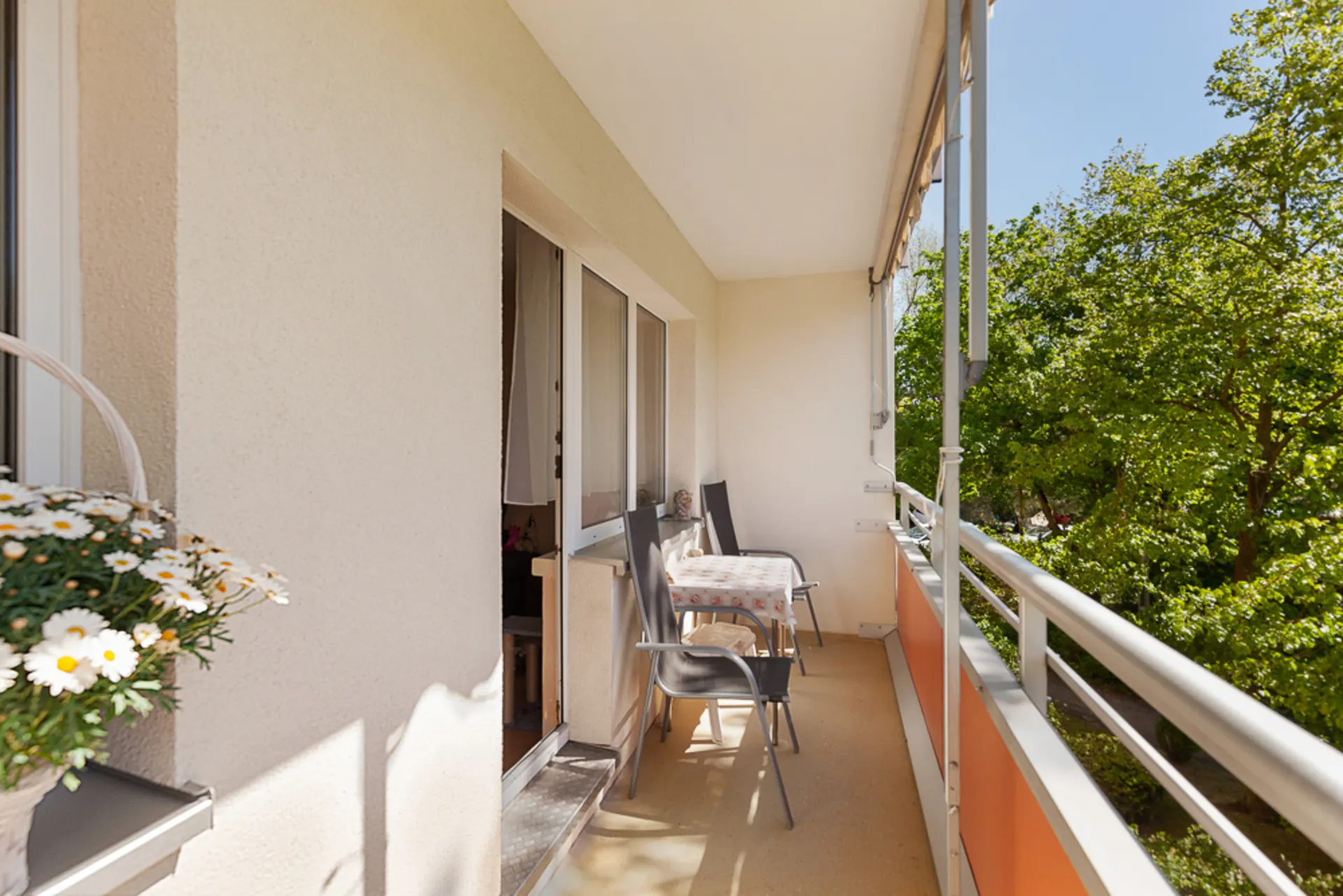 Balkon mit Tisch, zwei Stühlen und Blick auf grüne Bäume, Blumentopf links im Vordergrund.