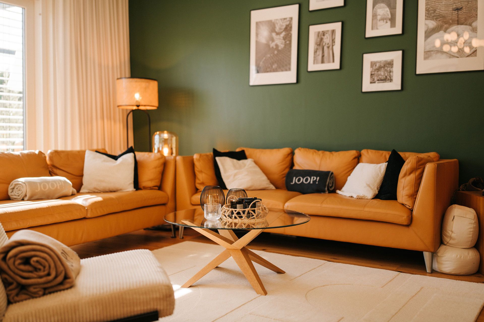 Wohnzimmer mit zwei gelben Sofas, Glastisch, Stehlampe und Bilderwand vor grüner Wand.