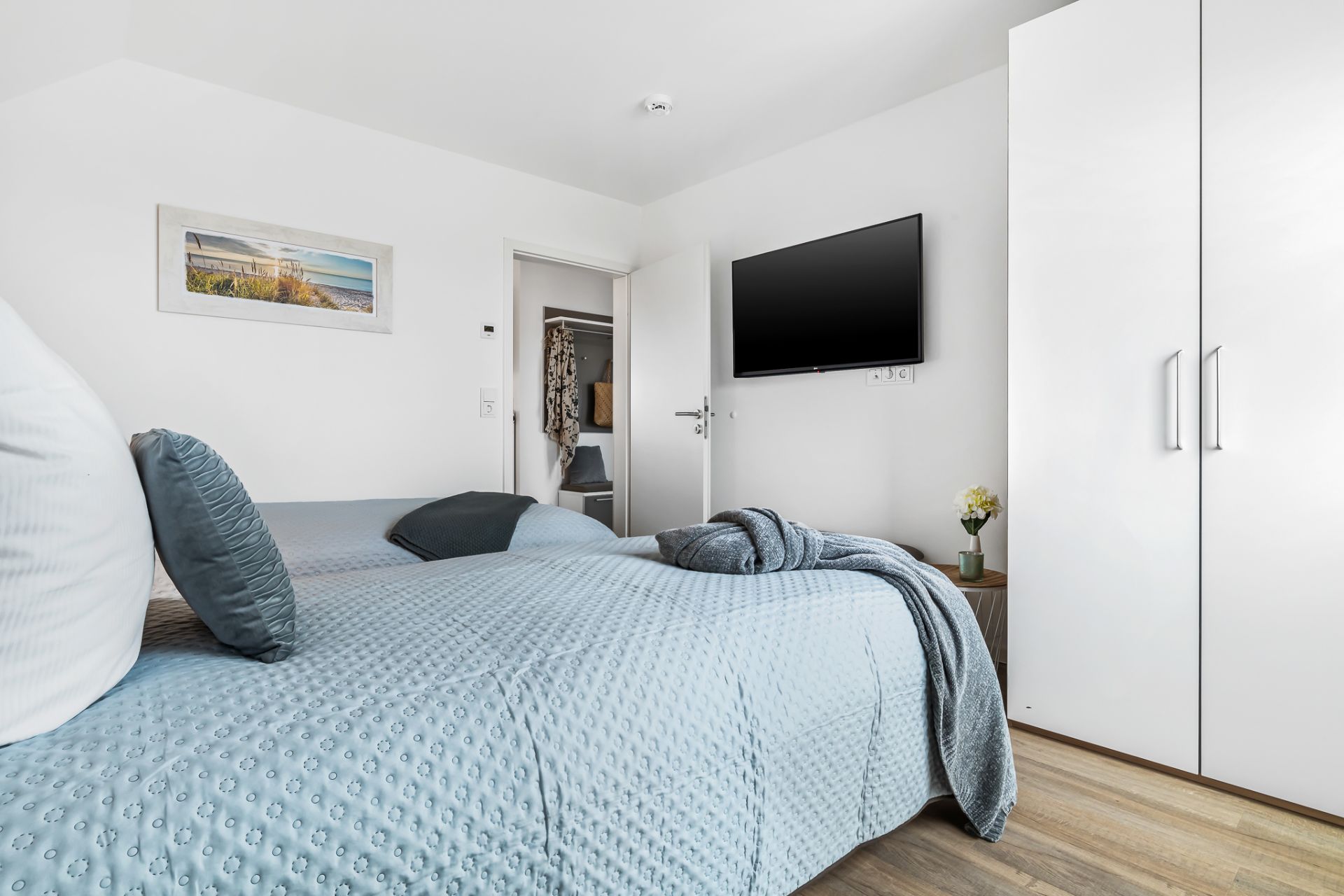Helles Schlafzimmer mit Doppelbett, Wandbild, TV, weißem Schrank und Nachttisch mit Blume.