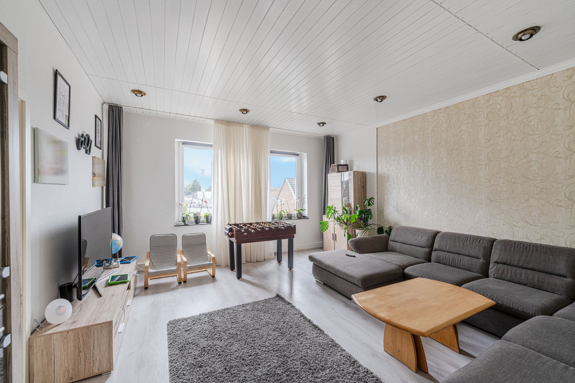 Helles Wohnzimmer mit grauem Sofa, Couchtisch, Fernseher, Kicker und großen Fenstern.