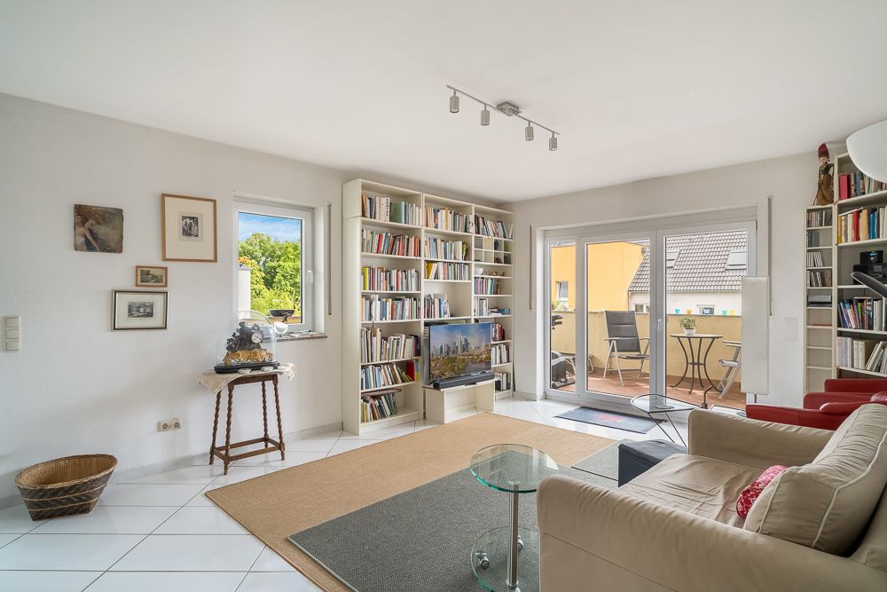 Helles Wohnzimmer mit Bücherregalen, Sofa, Fernseher und Zugang zu einem Balkon mit Tisch und Stühlen.