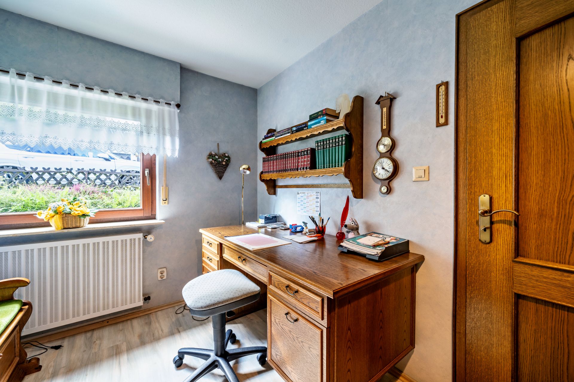Kleines Arbeitszimmer mit Holztisch, Regal, Stuhl und Fenster mit Gardine und Blumenkorb.