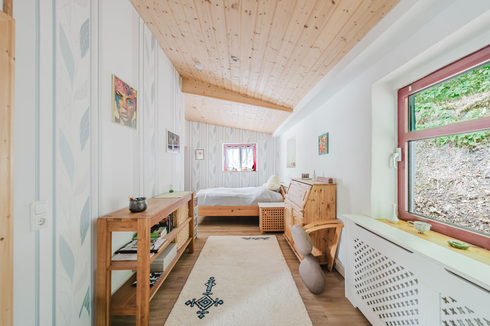 Helles Schlafzimmer mit Holzmöbeln, Teppich und großem Fenster mit Blick ins Grüne.