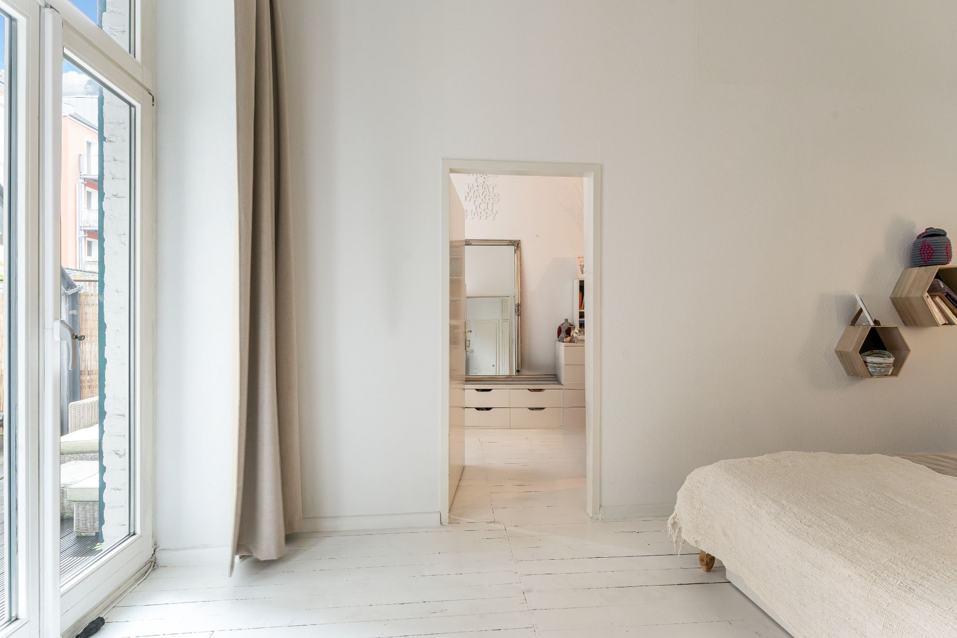 Helles Schlafzimmer mit Bett, Wandregalen und Blick auf einen Flur mit Kommode und Spiegel.