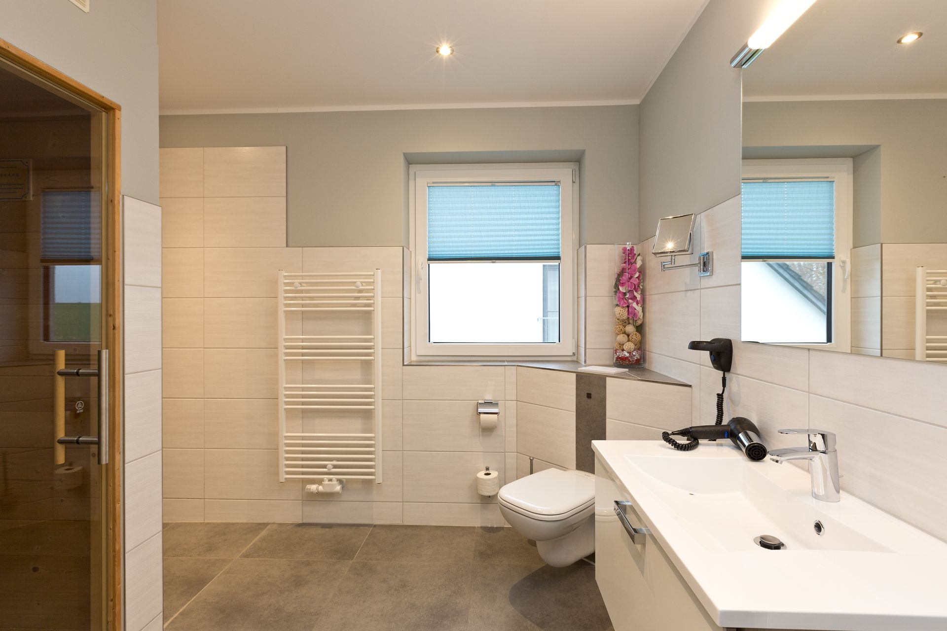Modernes Badezimmer mit Fenster, Waschbecken, WC, Spiegel und Handtuchheizkörper.
