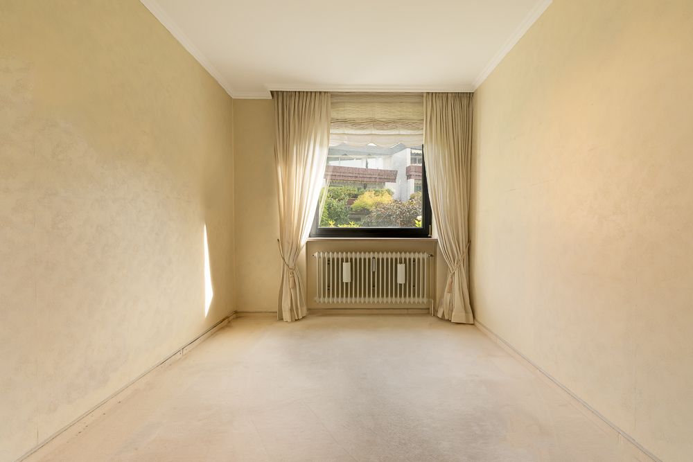 Leerer Raum mit beige Wänden, großem Fenster, Vorhängen und Heizkörper darunter.