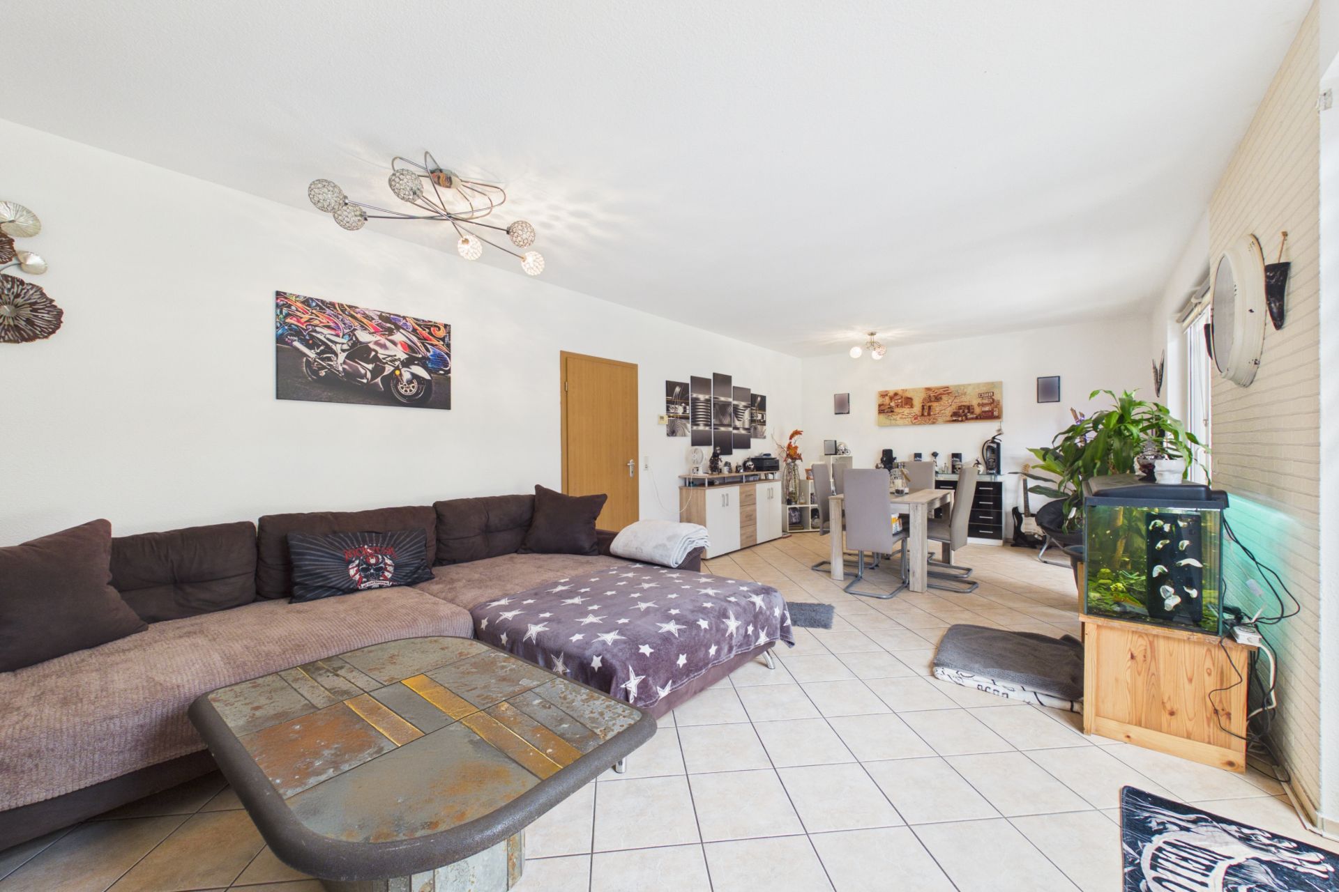 Wohnzimmer mit Ecksofa, Couchtisch, Esstisch, Aquarium und Wanddekorationen.
