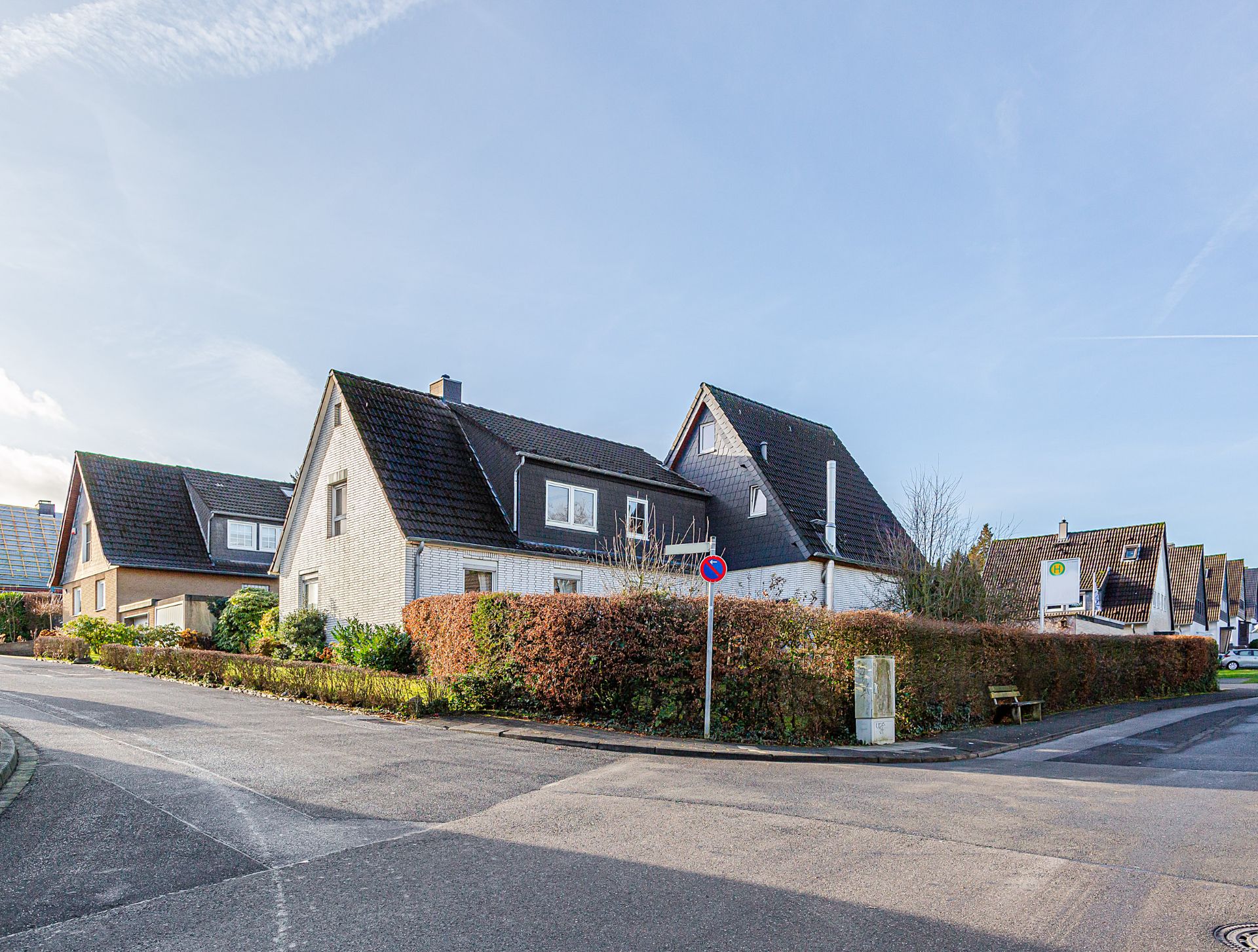 Einfamilienhaus mit schwarzem Dach an einer Straßenecke, Hecke und Bank im Vordergrund.