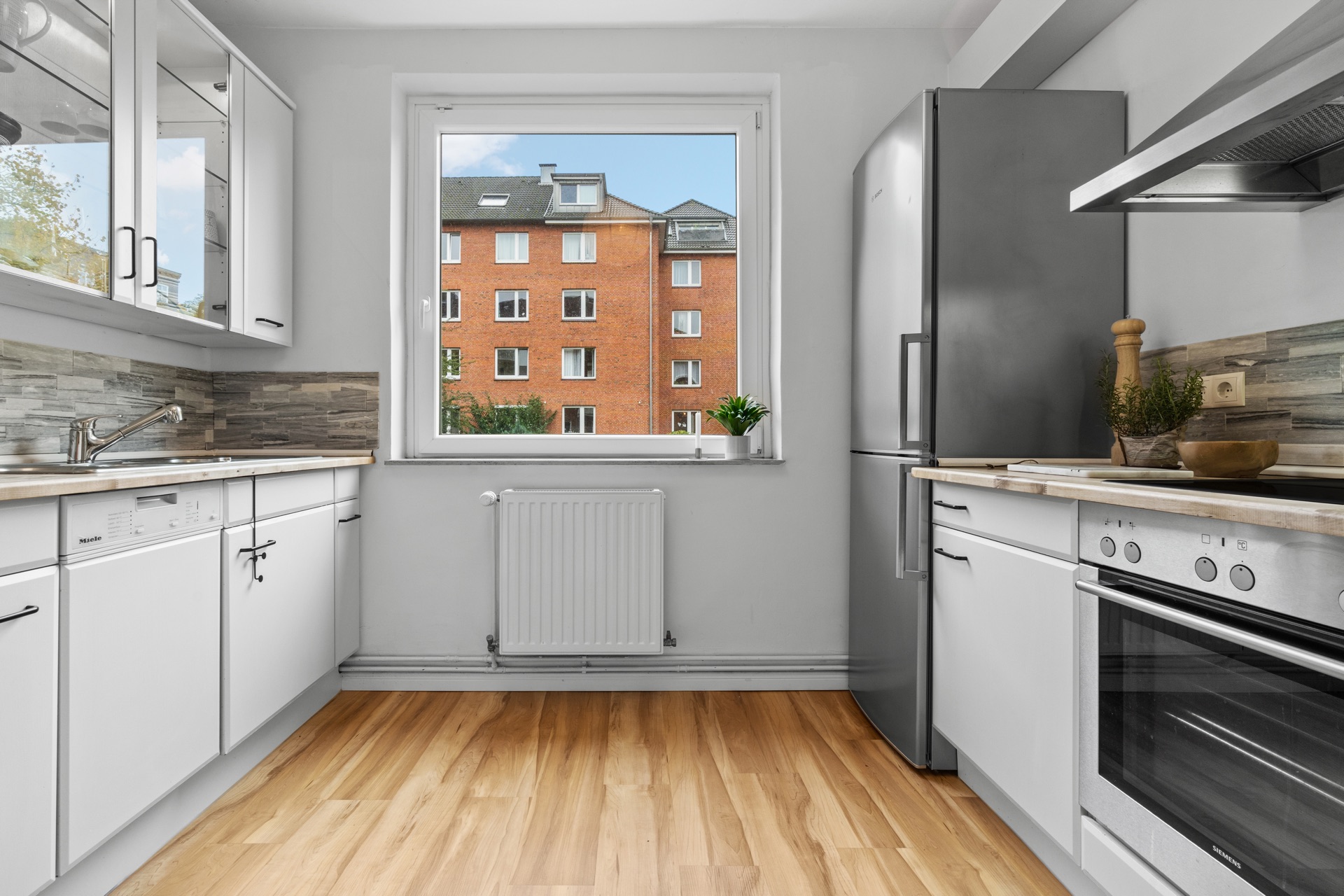 Moderne weiße Küche mit Holzfußboden, großem Fenster und Blick auf ein rotes Wohnhaus.