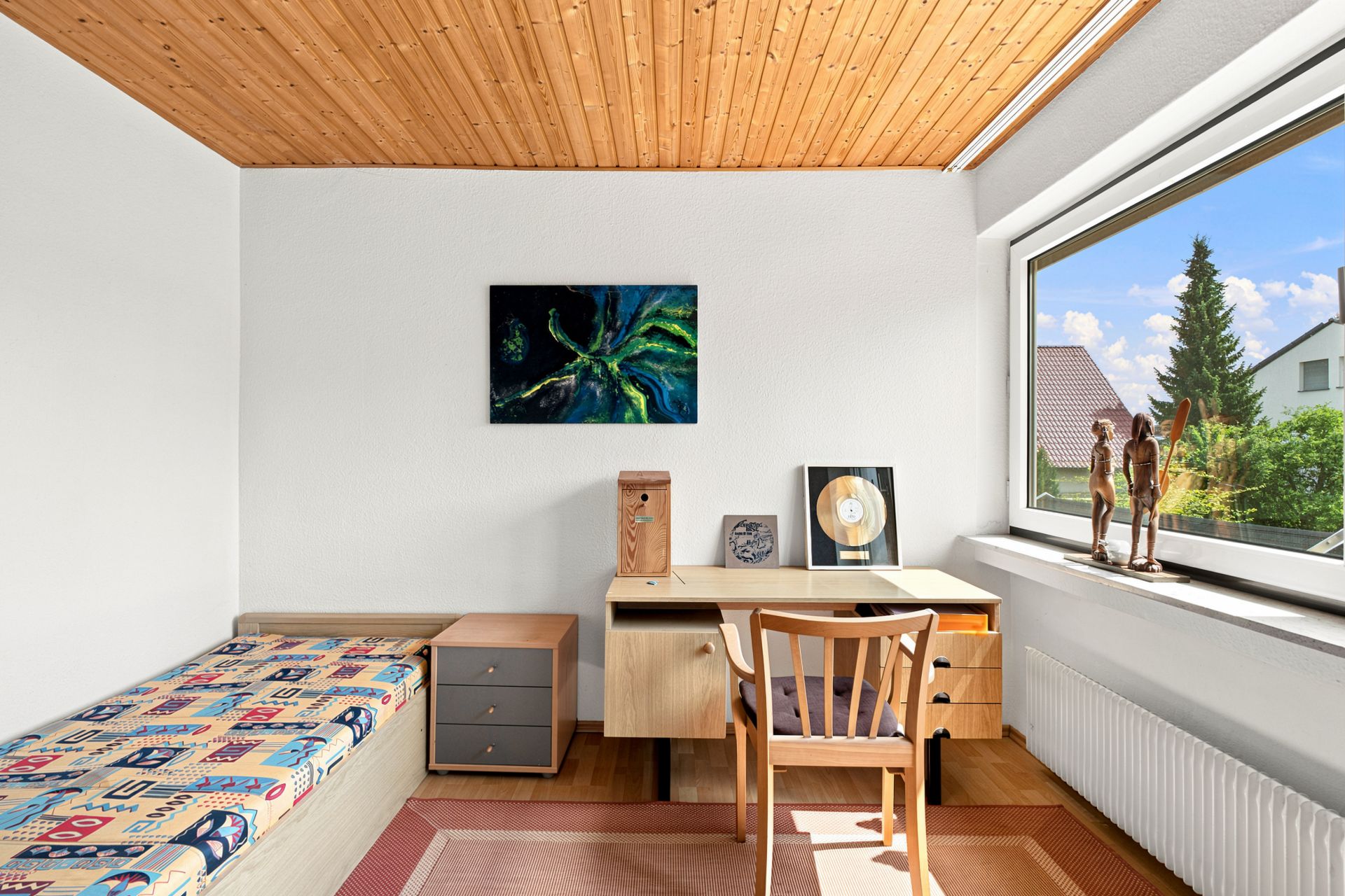 Helles Zimmer mit Einzelbett, Schreibtisch, Stuhl und großem Fenster mit Blick nach draußen.