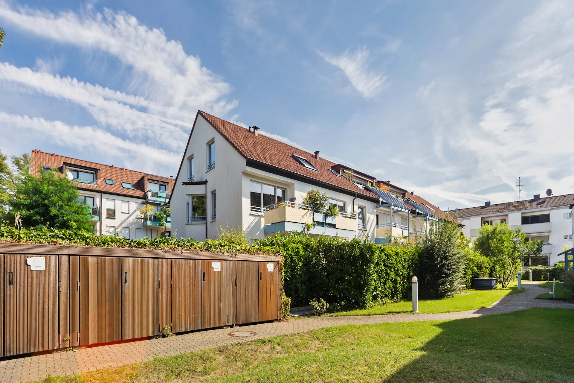 Mehrfamilienhaus mit Balkonen und Gartenanlage, Holzhütten im Vordergrund.