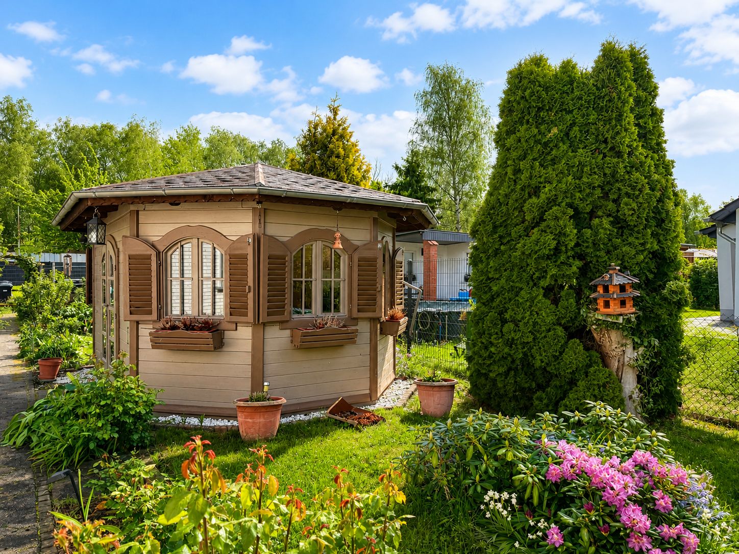 Kleines Gartenhaus mit braunen Fensterläden, umgeben von Pflanzen und Blumen.