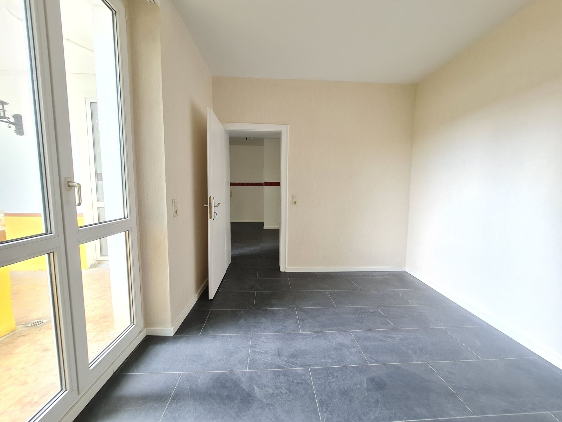 Helles Zimmer mit großen Fenstern, grauem Fliesenboden und geöffneter Tür zu einem weiteren Raum.
