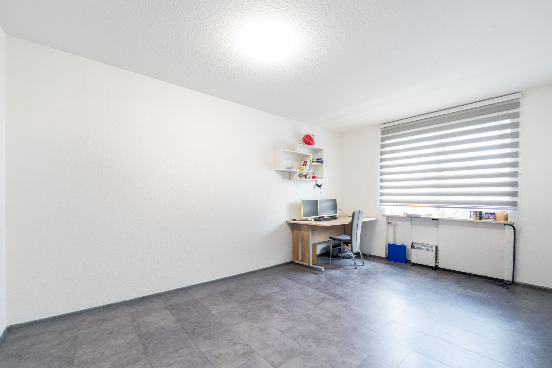 Helles Zimmer mit Schreibtisch, Stuhl, Regal und großem Fenster mit Jalousie.