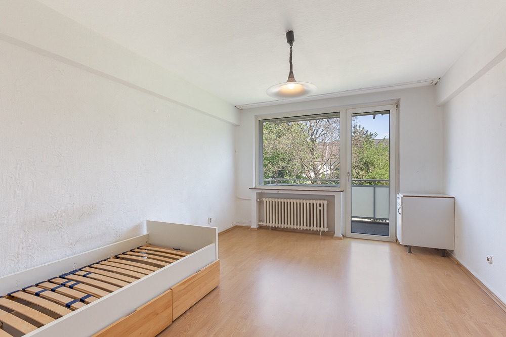 Helles Zimmer mit Holzboden, Einzelbettgestell, Fenster und Tür zu einem Balkon.