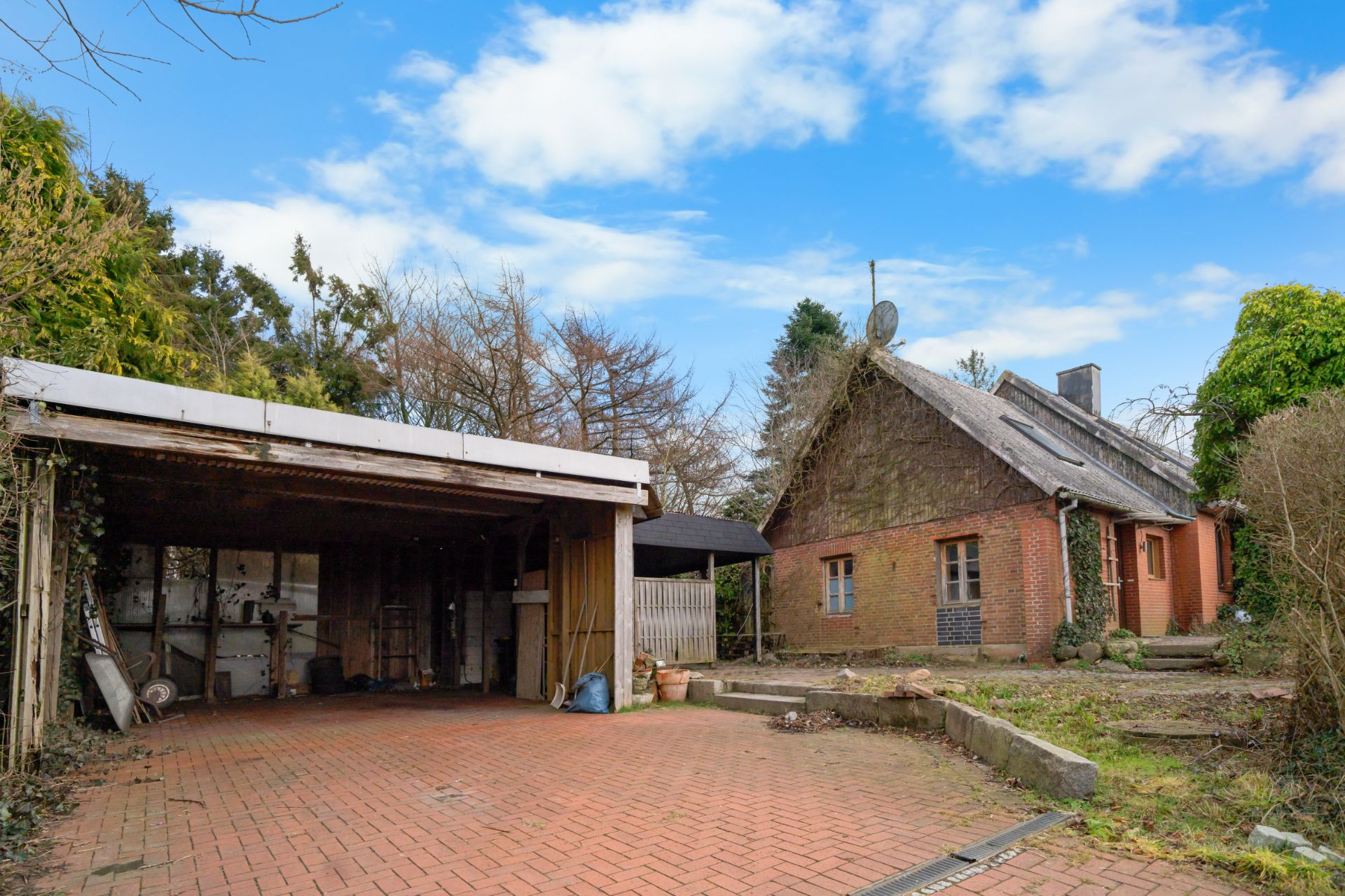 Backsteinwohnhaus mit Reetdach und offene Holzgarage auf gepflastertem Hof, umgeben von Bäumen.