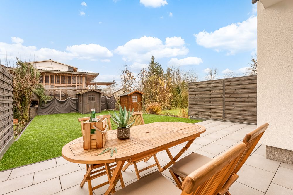 Holztisch mit Stühlen auf Terrasse, Blick auf Garten mit Rasen und zwei kleinen Holzhütten.