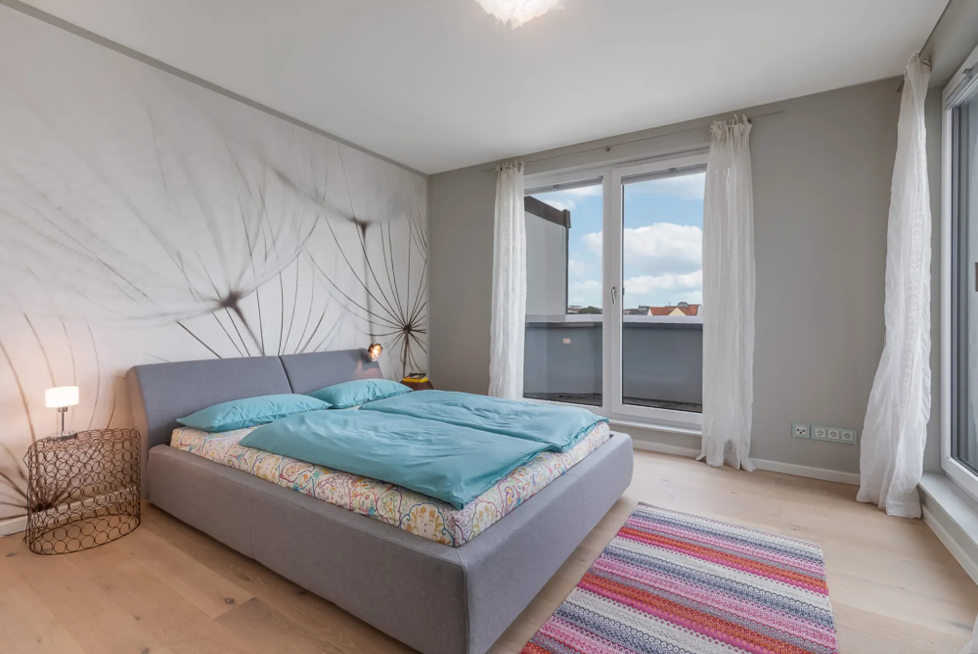 Helles Schlafzimmer mit großem Bett, Wandbild, buntem Teppich und Fenstern mit Blick nach draußen.