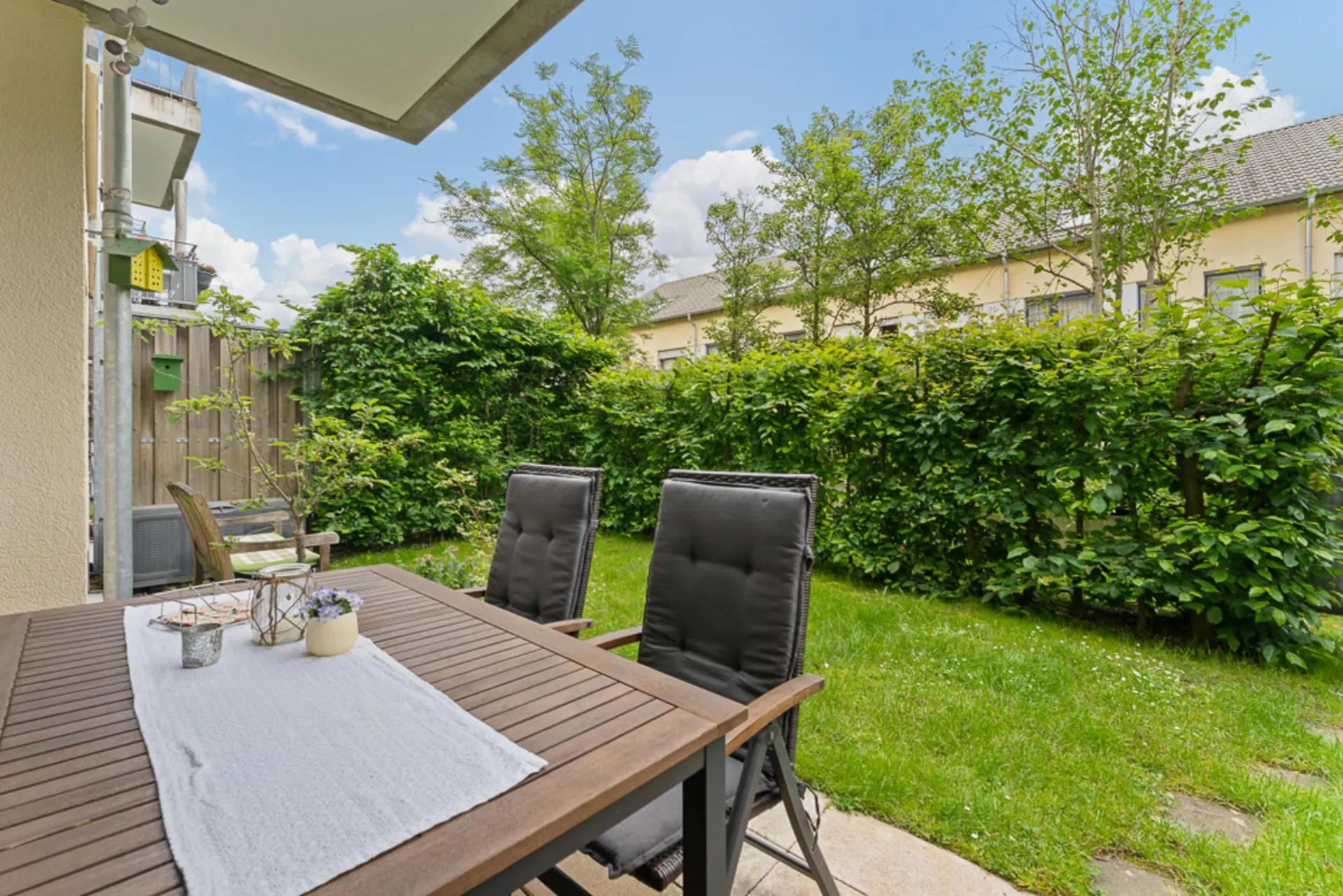 Terrasse mit Tisch und Stühlen, Blick auf grünen Garten mit Hecke und Bäumen.