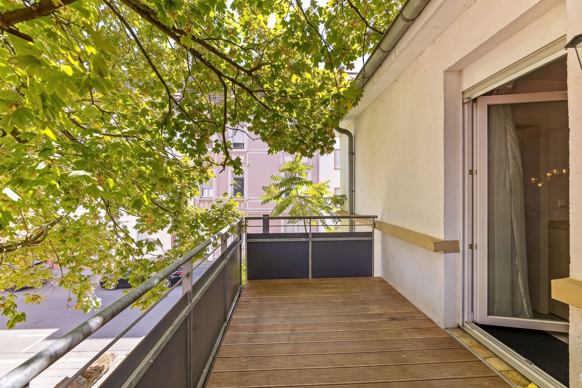 Holzbalkon mit Blick auf Bäume, angrenzend an ein weißes Wohnhaus mit geöffneter Glastür.