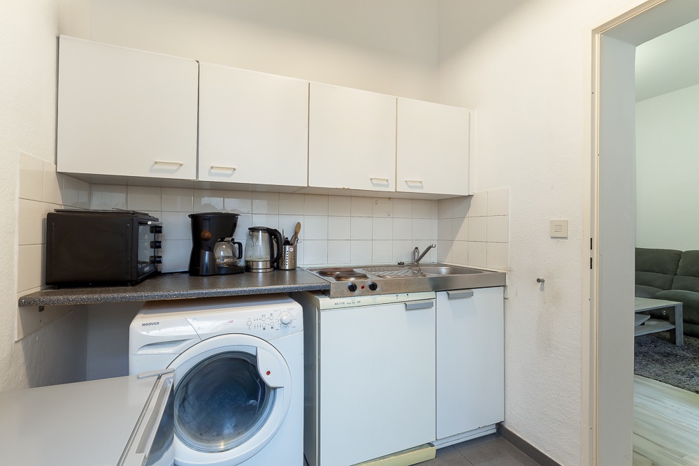 Kleine Küche mit Waschmaschine, Spüle, Mikrowelle, Kaffeemaschine, Wasserkocher und weißen Schränken.