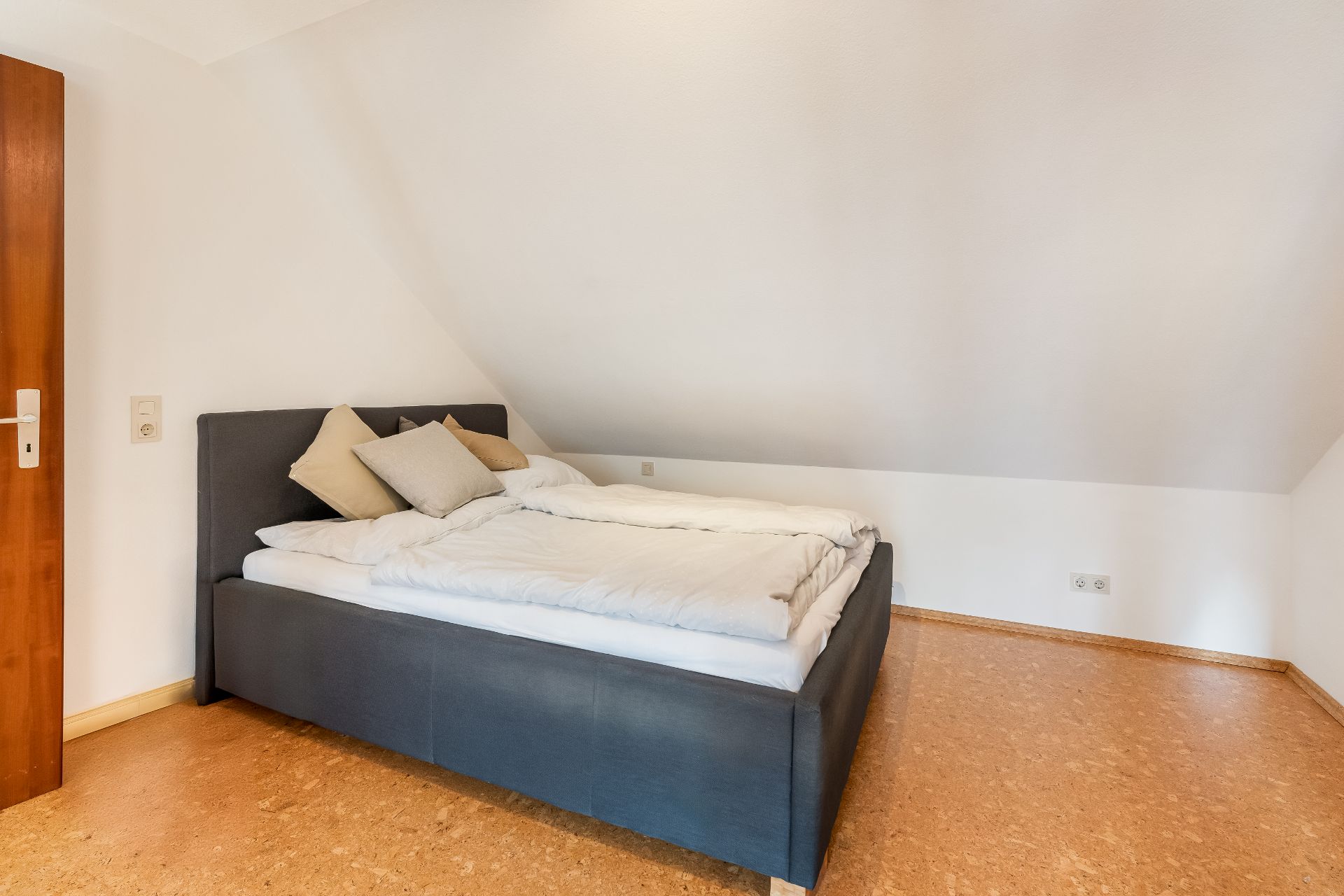 Schlafzimmer mit Doppelbett unter einer Dachschräge, Holzboden und weißen Wänden.
