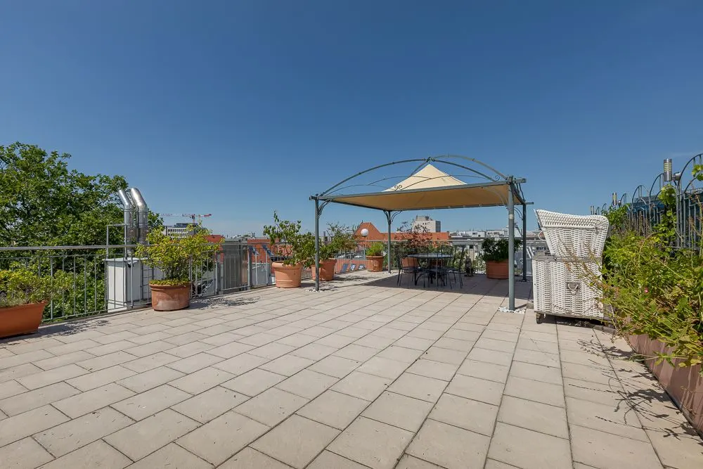 Große Dachterrasse mit Fliesen, Pflanzkübeln, Pavillon und Sitzbereich unter klarem Himmel.