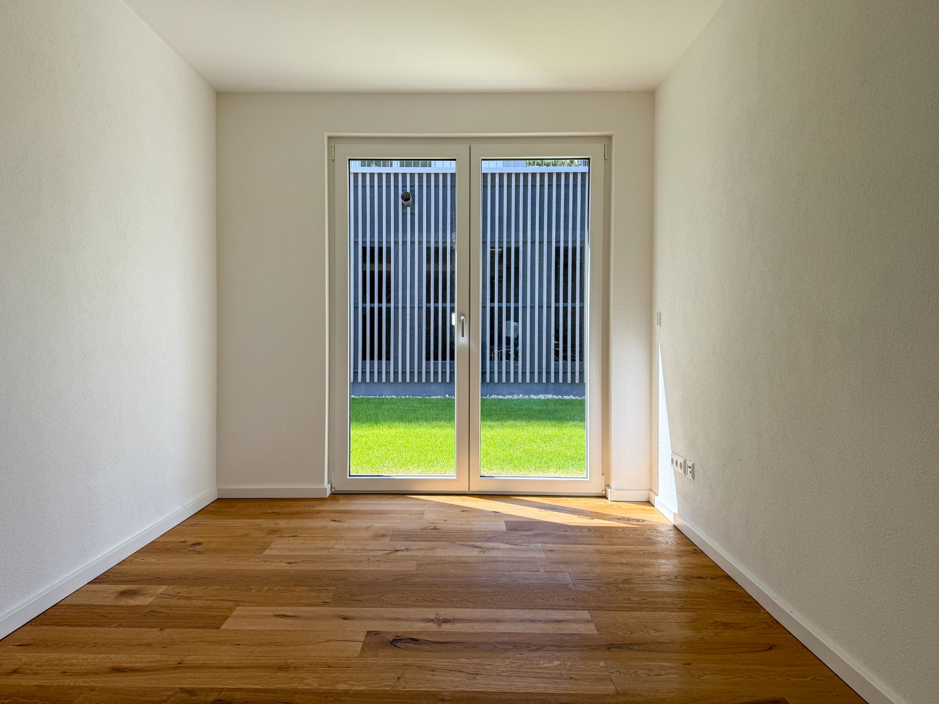 Leerer Raum mit Holzboden und bodentiefen Fenstern mit Blick auf Rasen.