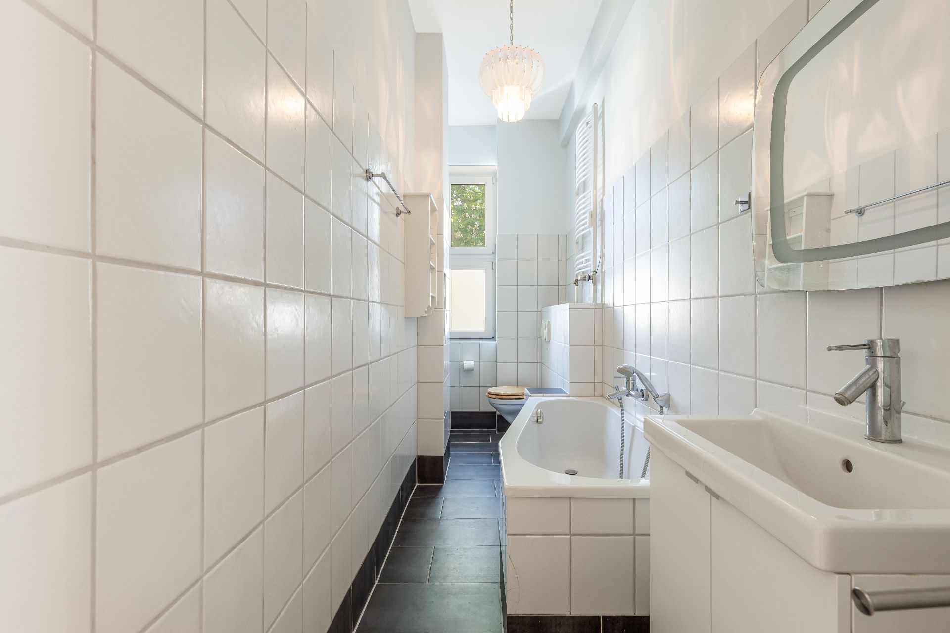Helles, schmales Badezimmer mit Badewanne, Waschbecken, Spiegel und Fenster.