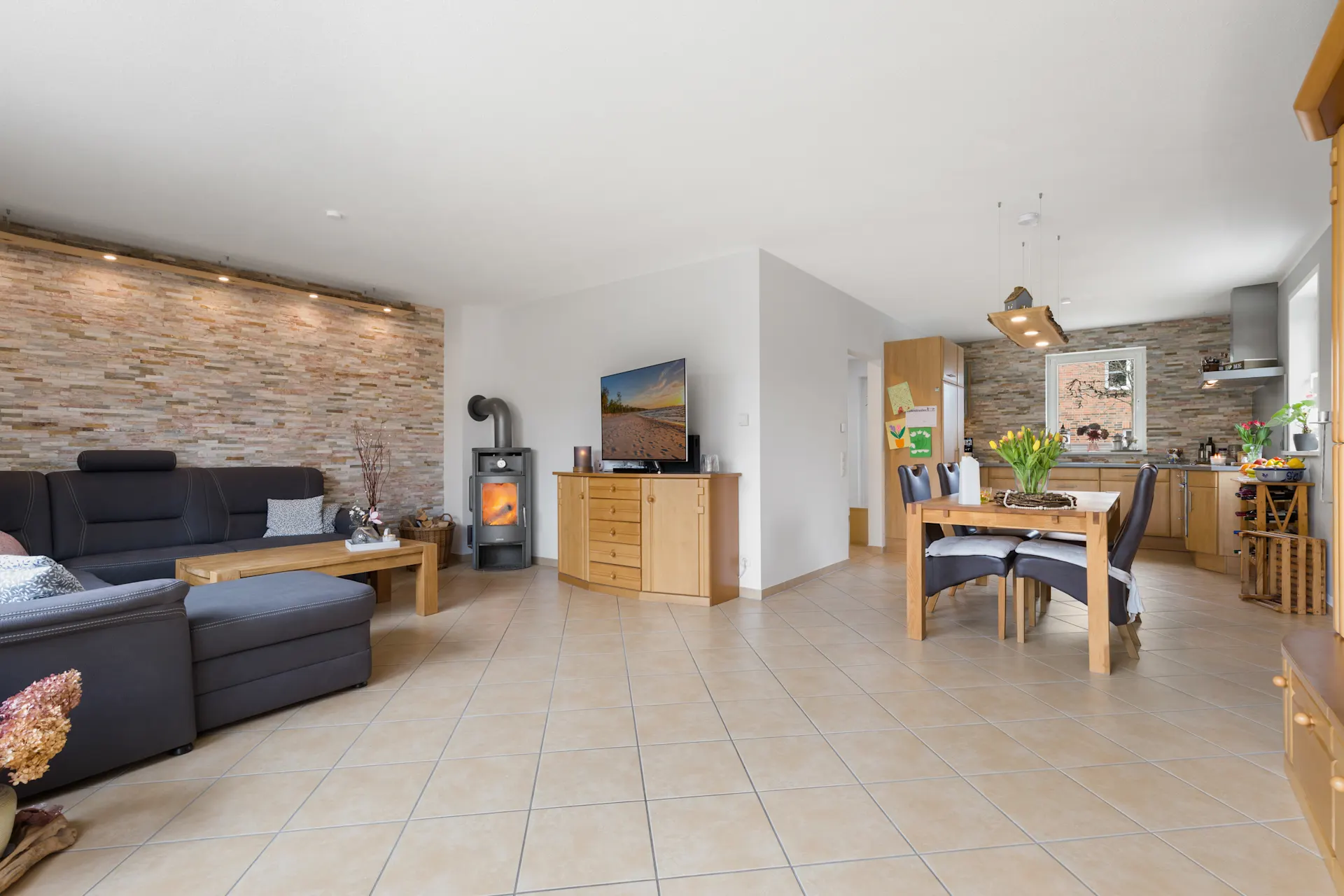 Helles Wohnzimmer mit Ecksofa, Holzofen, Essbereich und offener Küche, Fliesenboden.
