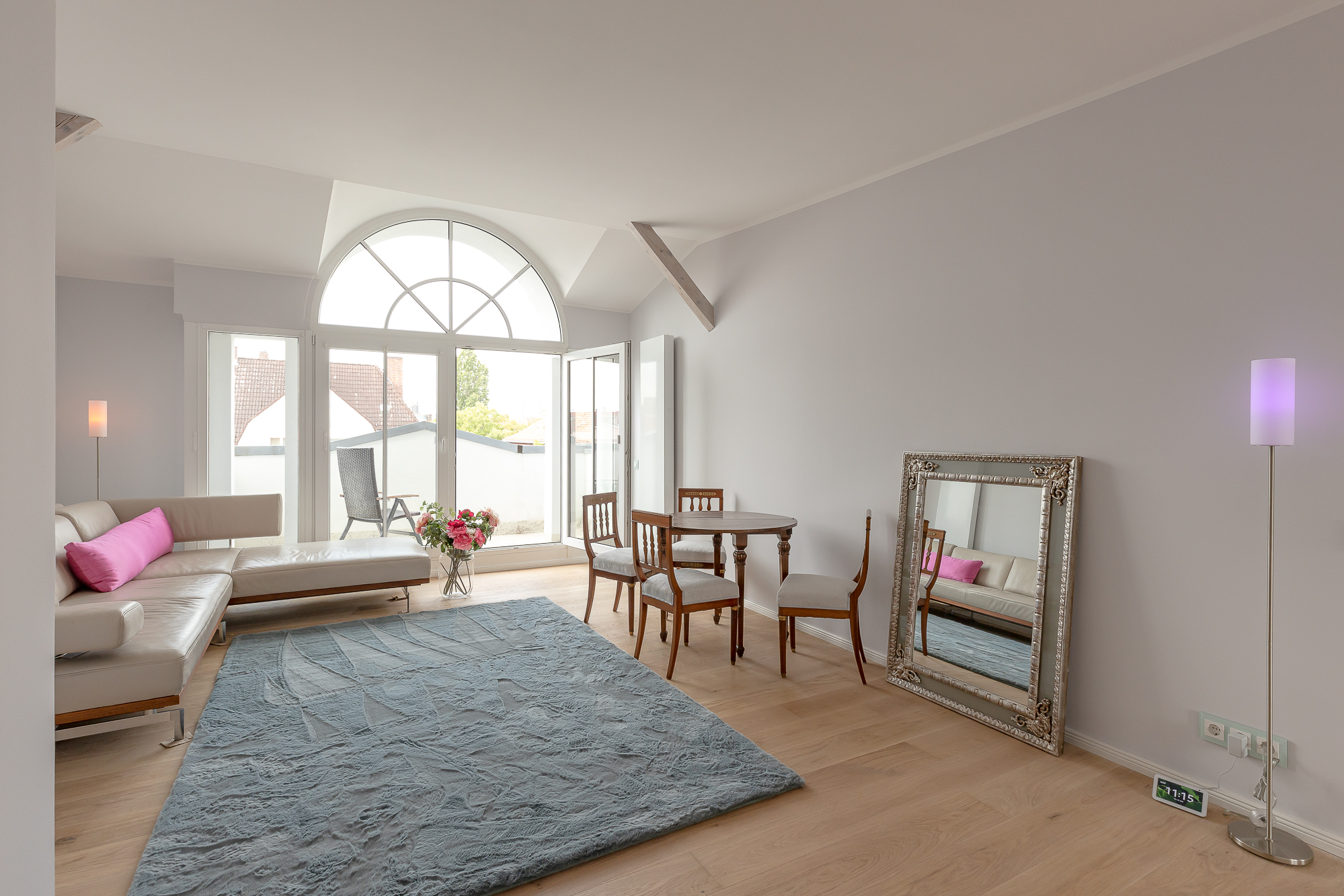 Helles Wohnzimmer mit Sofa, Esstisch, großem Spiegel, Teppich und bodentiefen Fenstern.