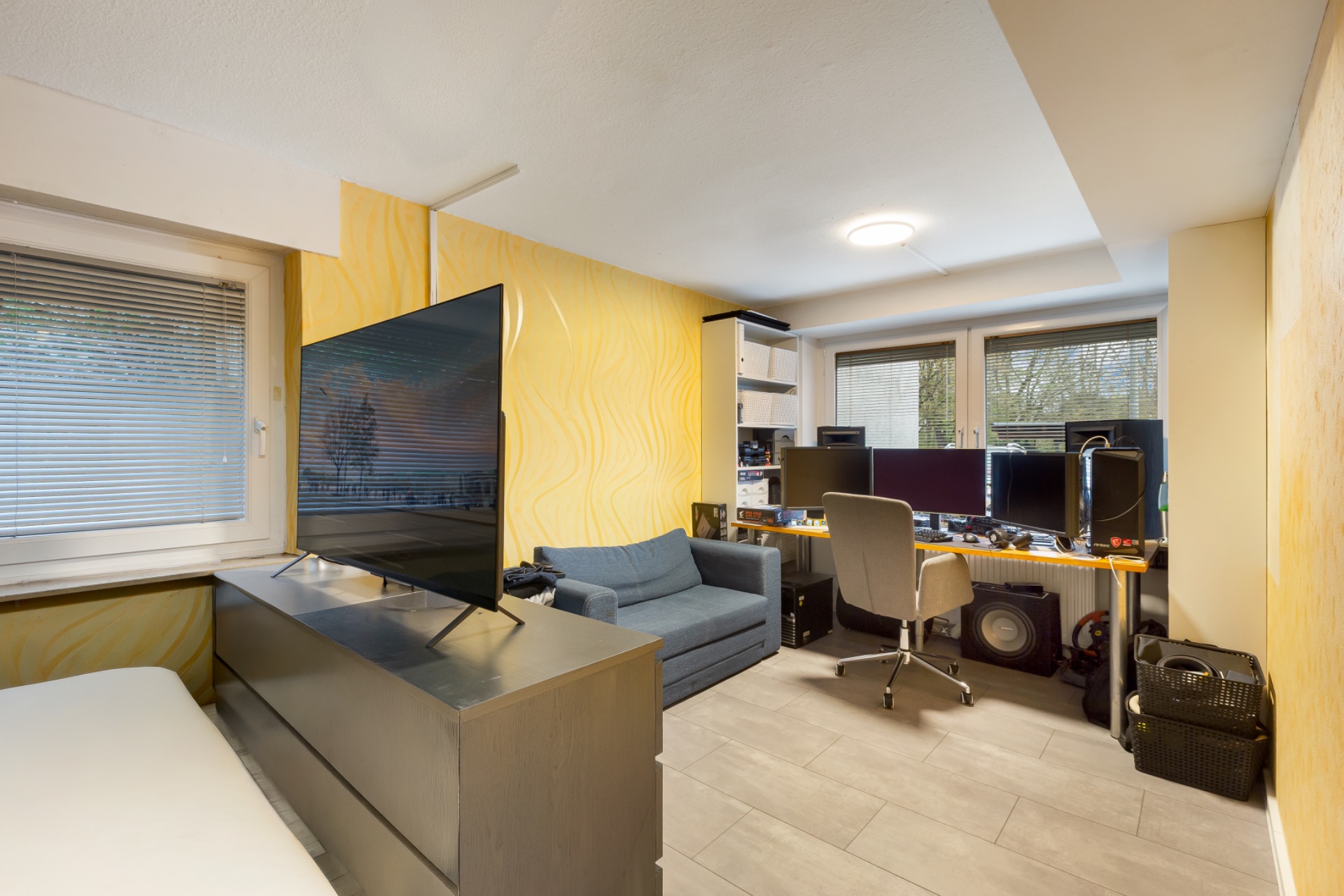Arbeitszimmer mit Schreibtisch, mehreren Monitoren, Sofa, Fernseher und großen Fenstern.