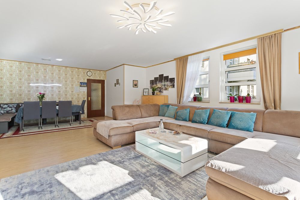 Helles Wohnzimmer mit großer Eckcouch, Glastisch, Esstisch und drei Fenstern mit Vorhängen.