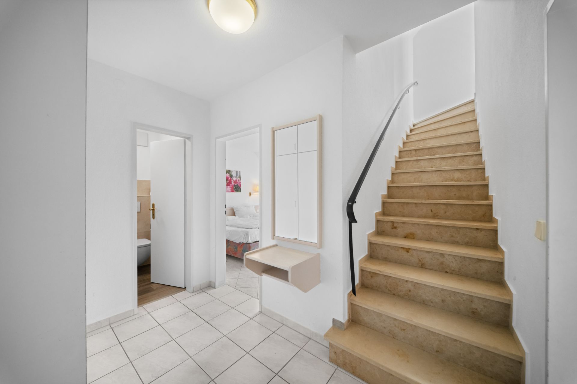 Flur mit Treppe nach oben, zwei offenen Türen und einem Wandspiegel mit Ablage.