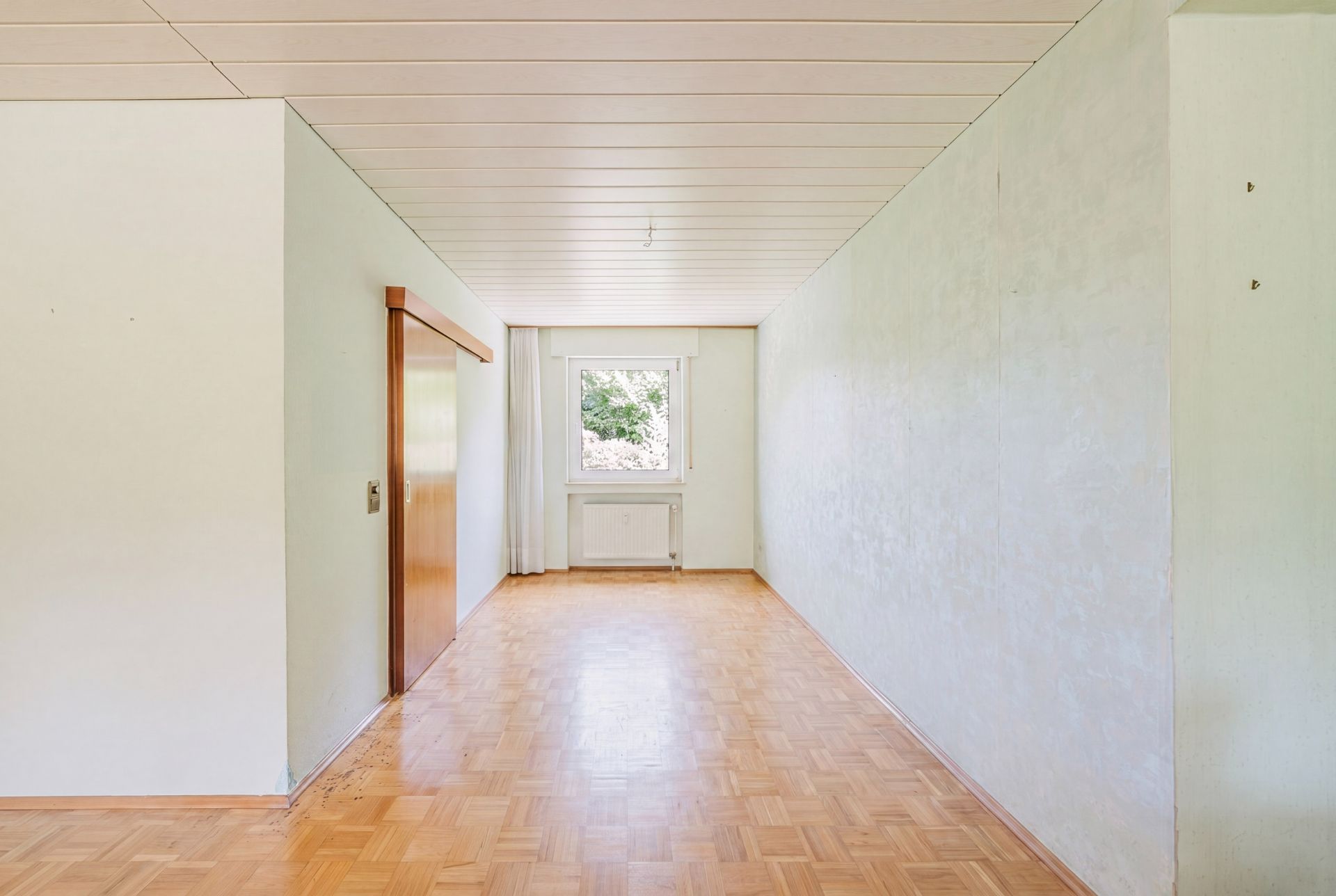 Leerer Raum mit Parkettboden, weißer Decke und einem Fenster am Ende.