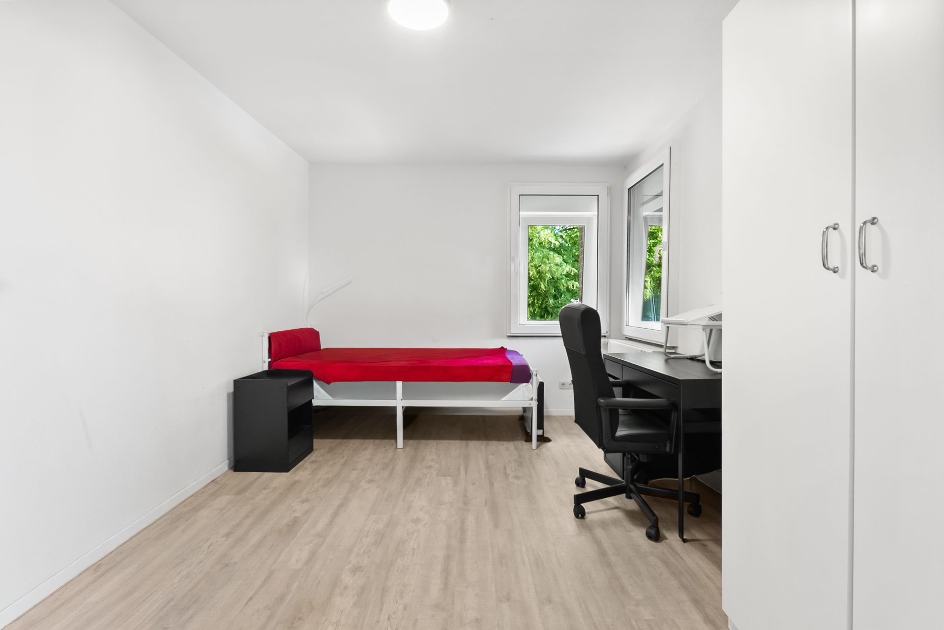 Helles Zimmer mit Einzelbett, Schreibtisch, Stuhl, Regal und Kleiderschrank auf Laminatboden.