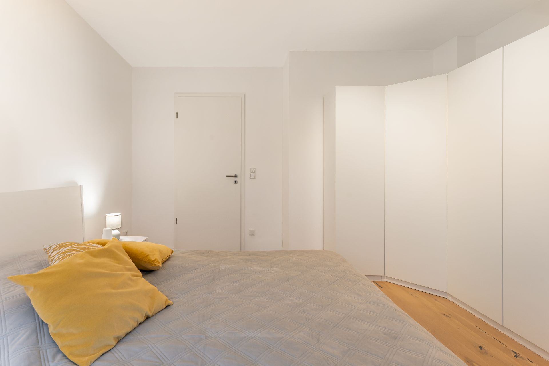 Helles Schlafzimmer mit großem Bett, gelben Kissen, Nachttisch und weißem Kleiderschrank.