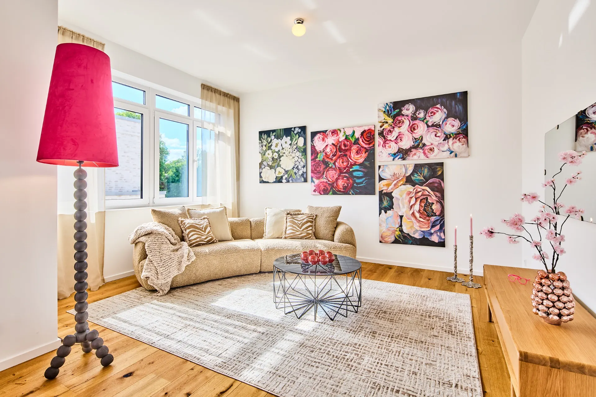 Helles Wohnzimmer mit beige Sofa, runder Glastisch, pinker Stehlampe und Blumenbildern an der Wand.