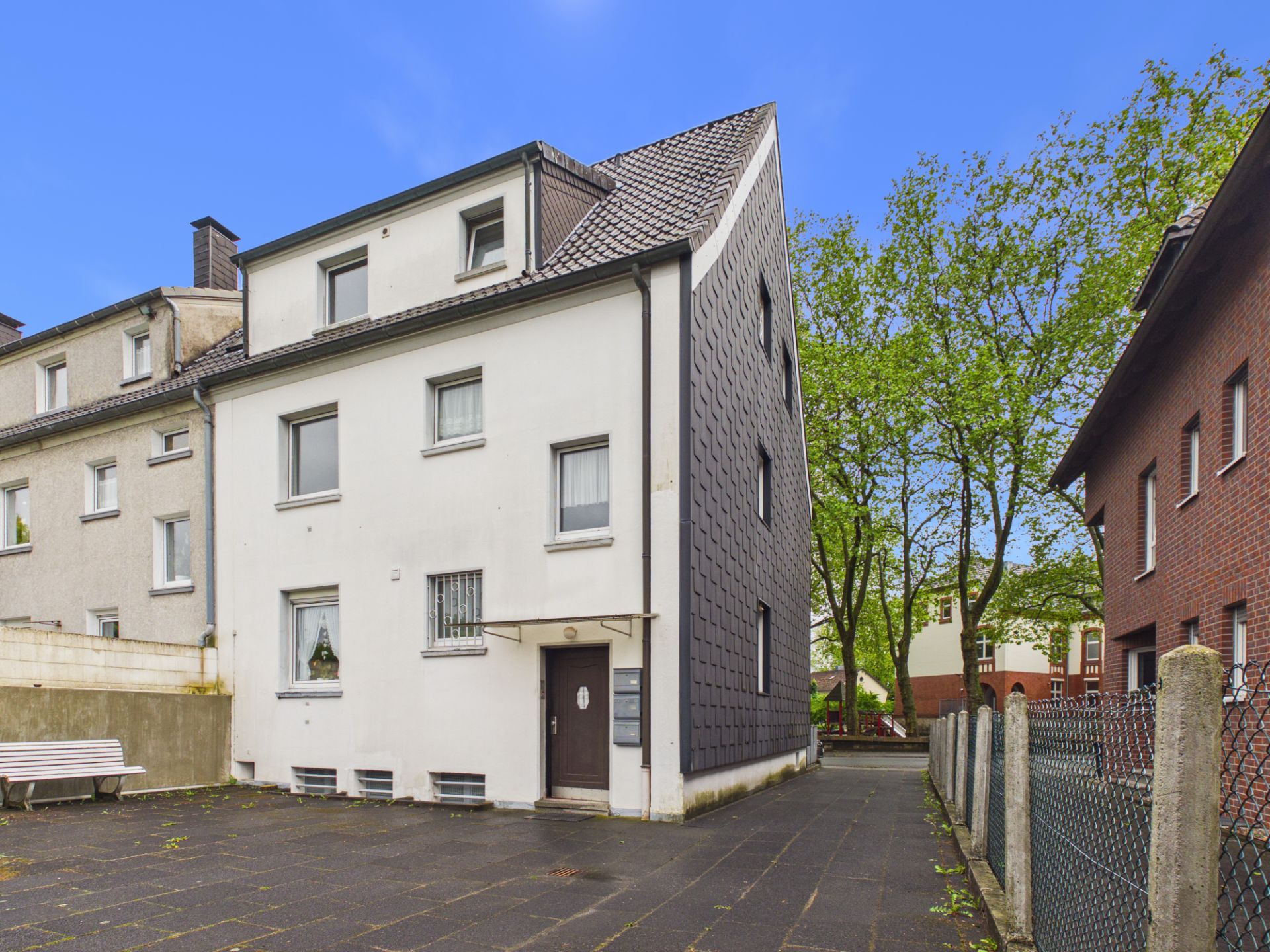 Dreistöckiges Wohnhaus mit weißer Fassade und schrägem Dach, daneben eine Bank und Bäume im Hintergrund.