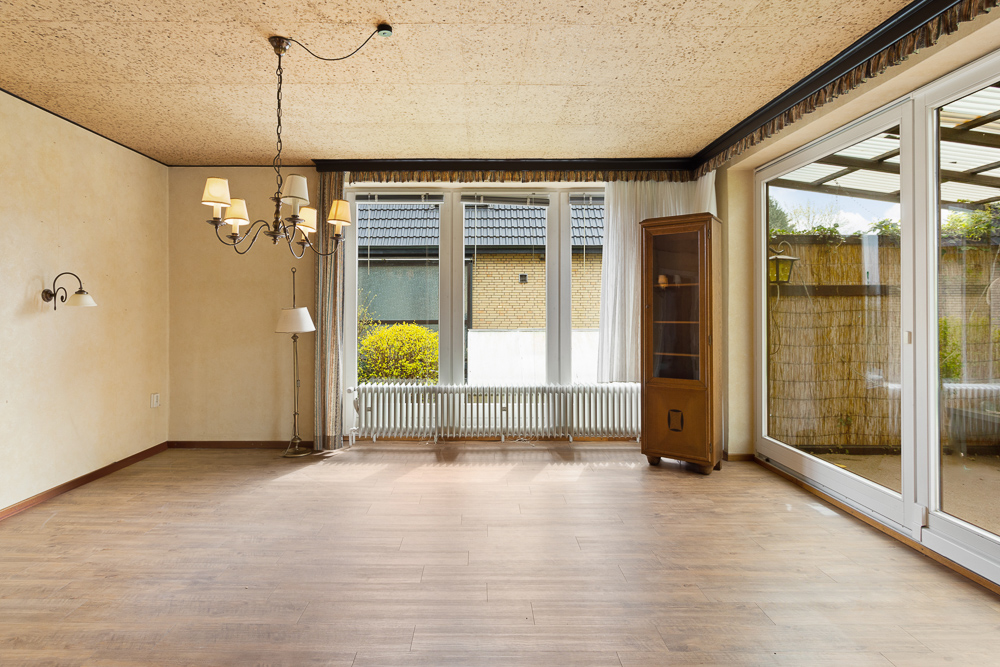 Leeres Wohnzimmer mit Holzfußboden, Kronleuchter, Stehlampe, Glasschrank und großen Fenstern.