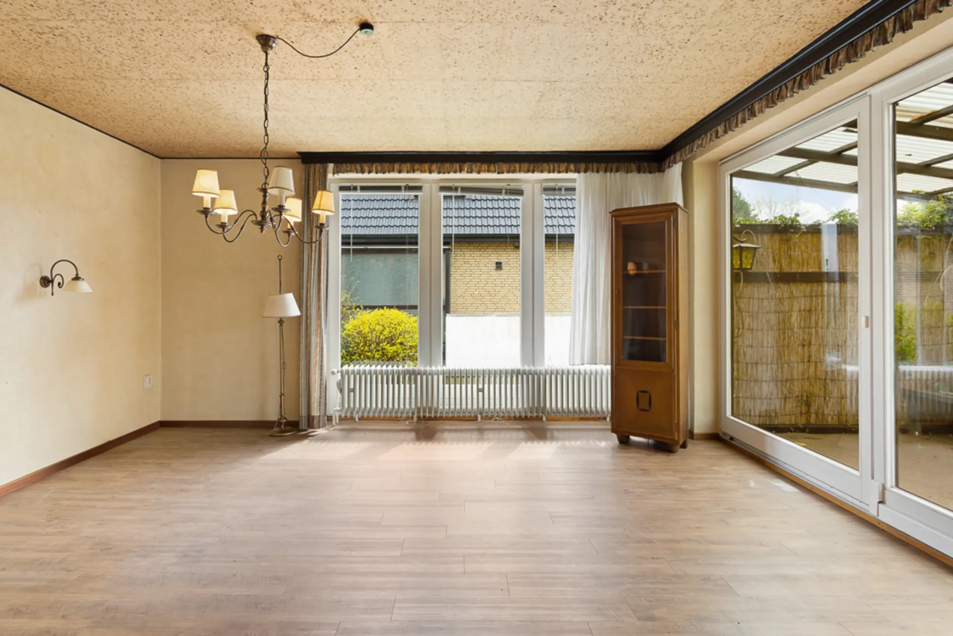 Leeres Wohnzimmer mit Holzfußboden, Kronleuchter, Stehlampe, Glasschrank und großen Fenstern.